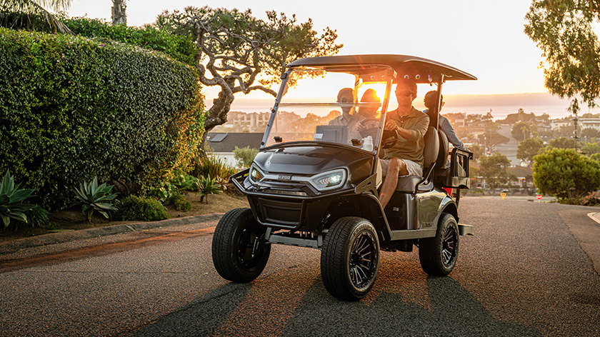 E-Z-GO Express 4 driving in a neighborhood with city, sunset, shrubs, and trees in background